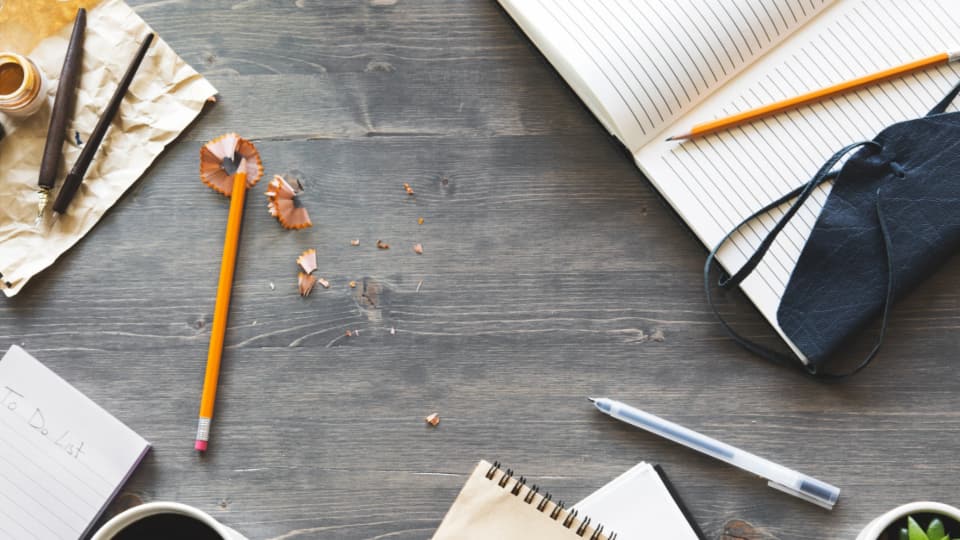 A dark wooden desk seen from above, with a blank to-do list pad, a sharpened pencil, pencil shavings, an open notebook and a cup of coffee.