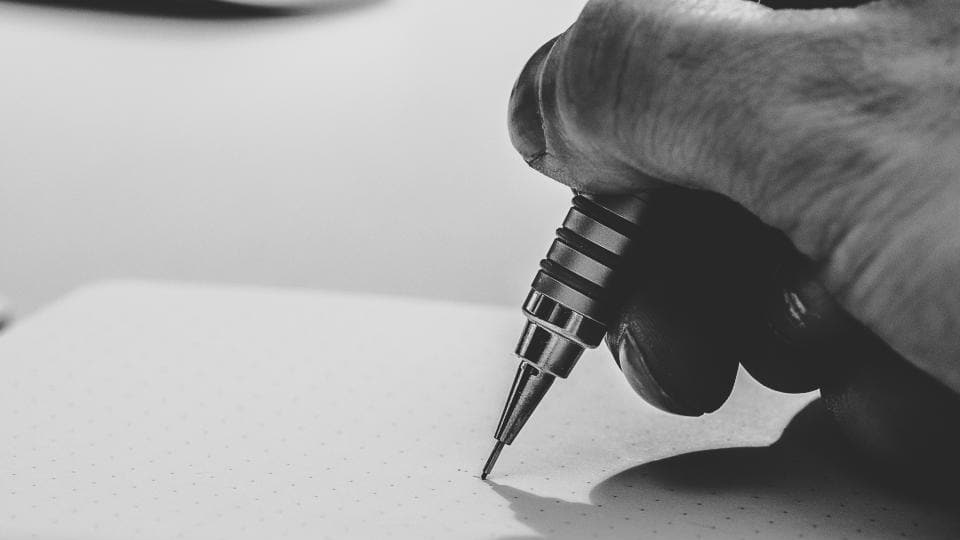 A close-up in black and white of a hand holding a mechanical pencil, its tip just touching a blank dotted notebook page.