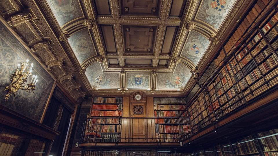 The interior of a grand old library, shot looking upwards at a painted and gilded ceiling above two storeys of full bookshelves.